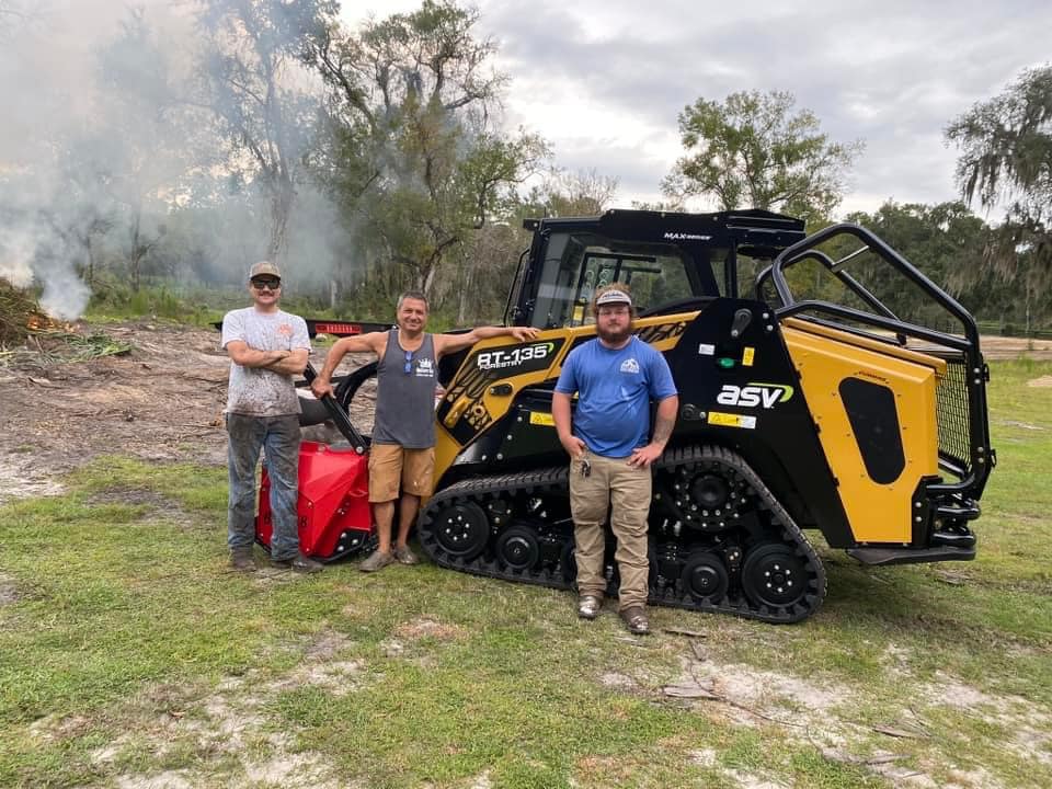 Three men stand beside a tractor and a brush cutter, showcasing agricultural equipment in an outdoor setting.